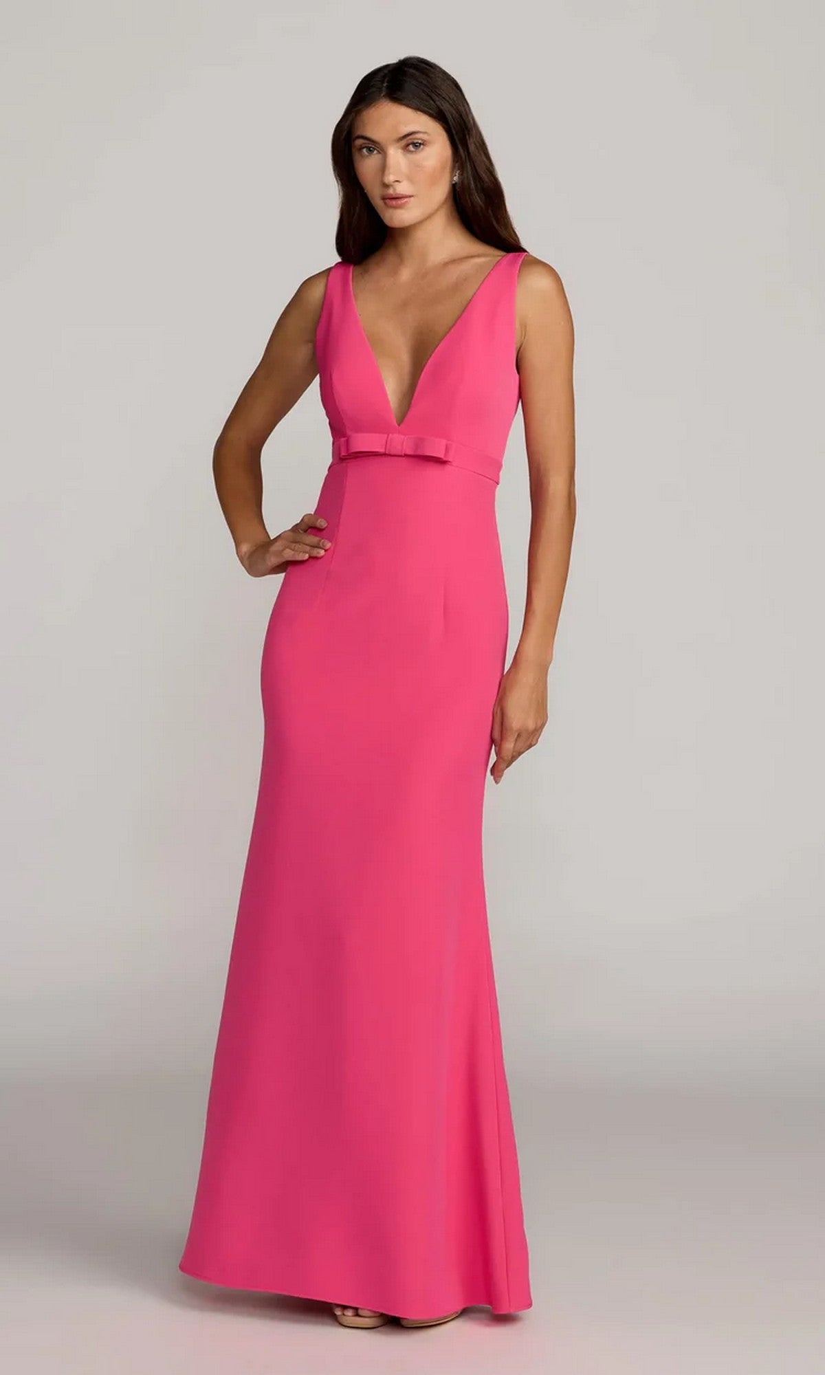 A woman stands against a light gray background, wearing the Mac Duggal 2371 Long Formal Dress—a bright pink, sleeveless crepe gown with a deep V-neckline and small waist bow.